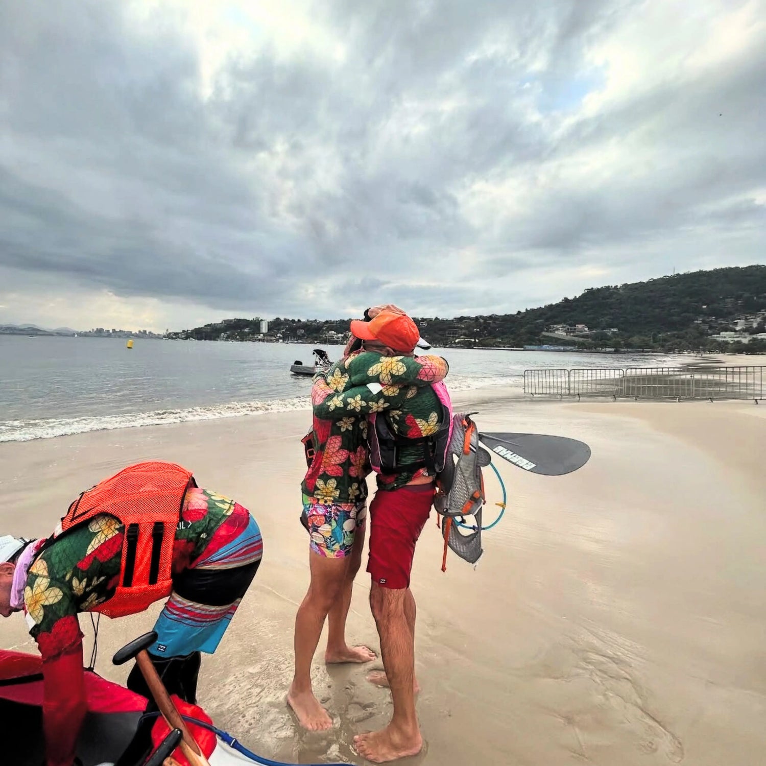 Two athletes in floral-patterned performance gear from the Champion collection embrace on a beach next to their racing canoe after a successful competition