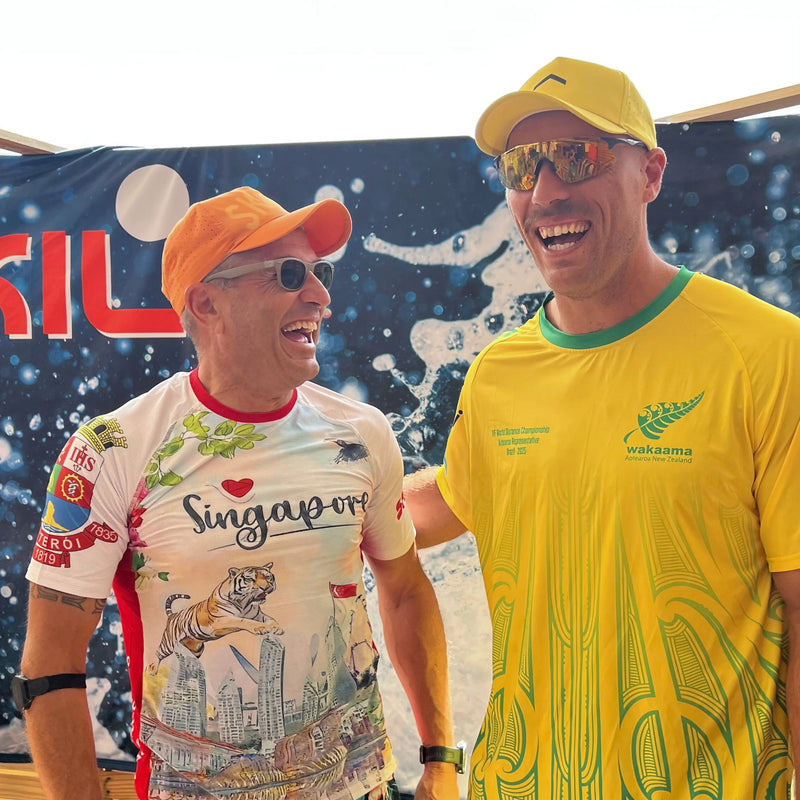 Two male athletes celebrate at the IVF World Distance Championships, one wearing a custom Singapore-themed performance rashguard from the Champion collection and the other in a New Zealand Waka Ama jersey.