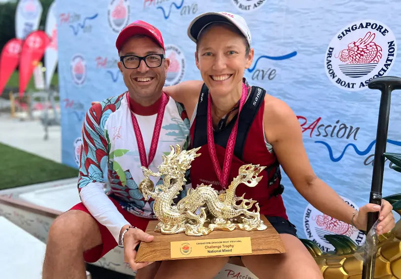 Two smiling athletes hold a gold dragon trophy at the Singapore Dragon Boat Festival, with one wearing a long-sleeve rashguard from the Japanese Wings collection featuring red and blue wing patterns.