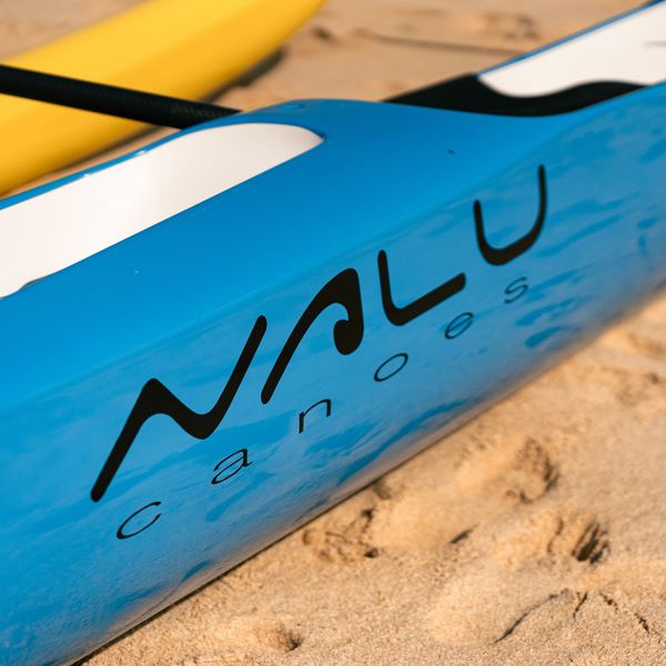 A technical close-up of a light blue Synergy tandem racing canoe from the Nalu collection, showing the "NALU canoes" branding on the side of the hull against the sand.