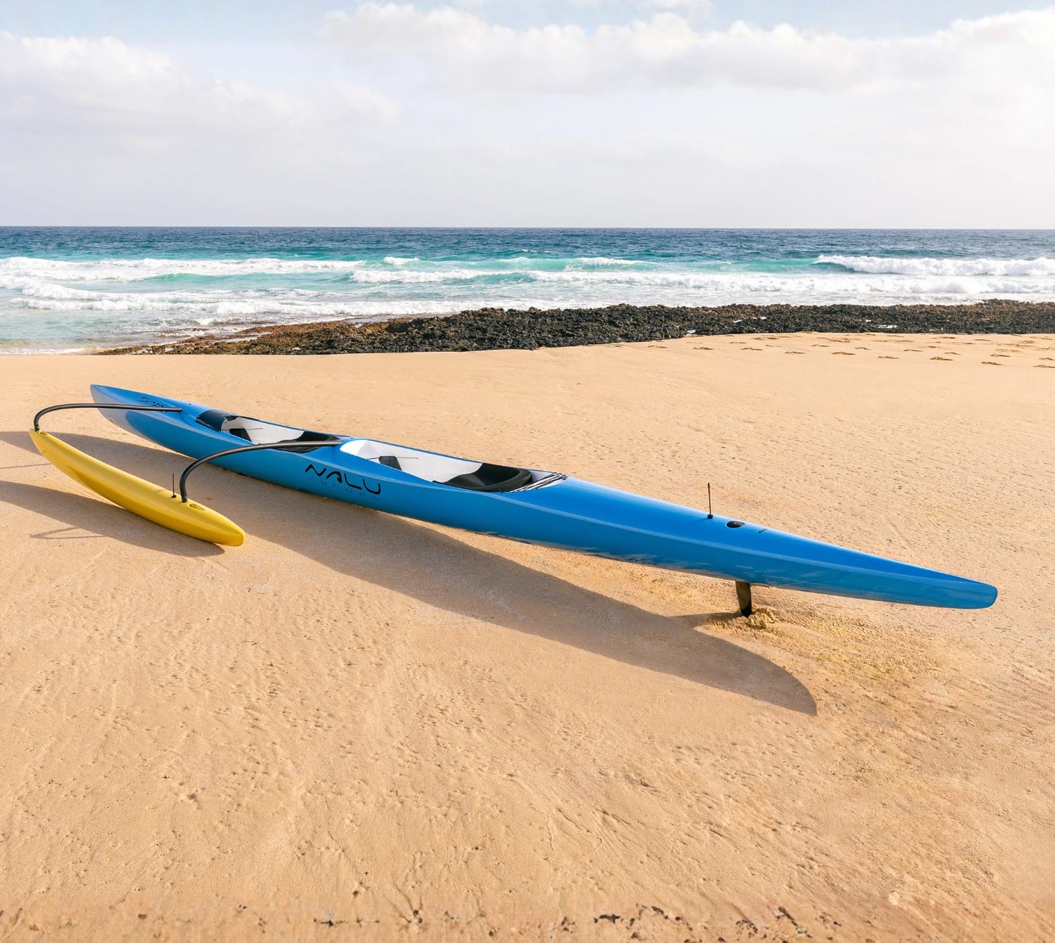 A sleek blue racing canoe with a yellow outrigger from the Nalu Outrigger collection rests on a sandy beach under a light blue sky.