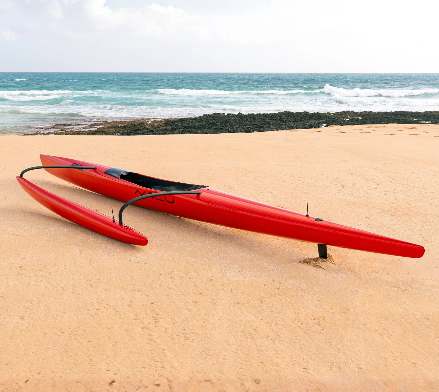 A professional red racing canoe from the Nalu Outrigger collection rests on a sandy beach, showcasing its aerodynamic design and carbon-fiber construction against a backdrop of ocean waves.