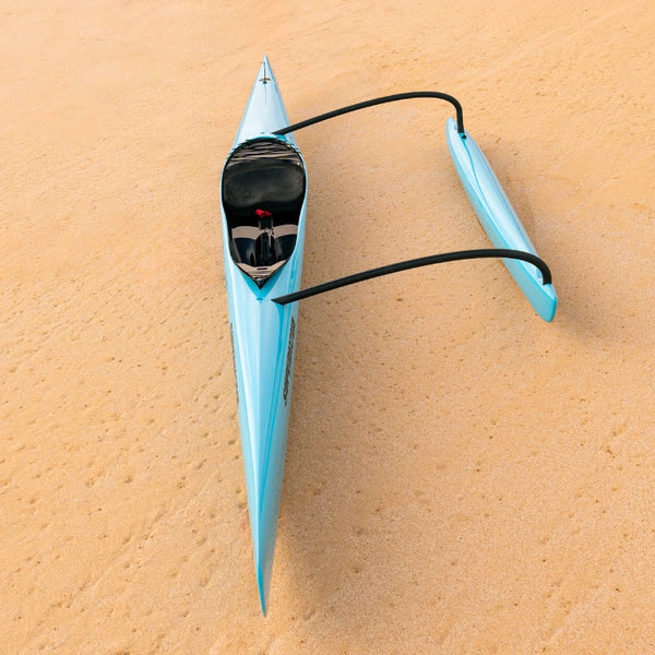 An overhead full-length shot of a light blue Supernova outrigger canoe with a black ama and connecting iakos, resting on golden sand.