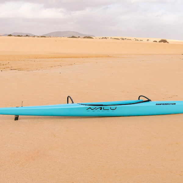 A sleek teal hull of a Supernova High Performance Canoe Collection canoe resting on a vast sandy beach with distant dunes, highlighting the vibrant Nalu branding.