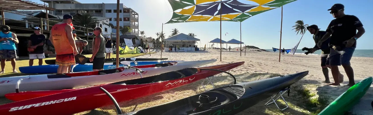 A panoramic view of professional Nalu outrigger canoes, including the Supernova model, lined up on a sunny beach under a geometric sunshade during a racing event.