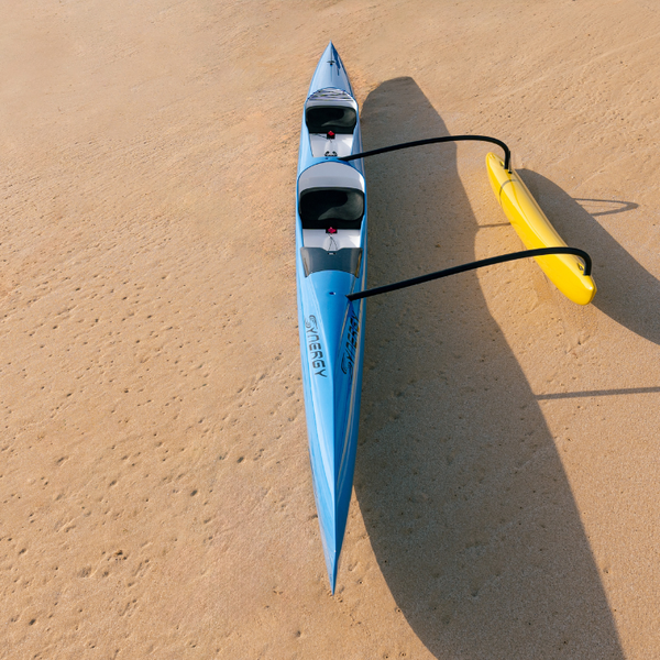 An overhead view of a sleek light blue Synergy double outrigger canoe from the Nalu collection resting on a sandy beach, featuring a high-visibility yellow ama and dual ergonomic cockpits.