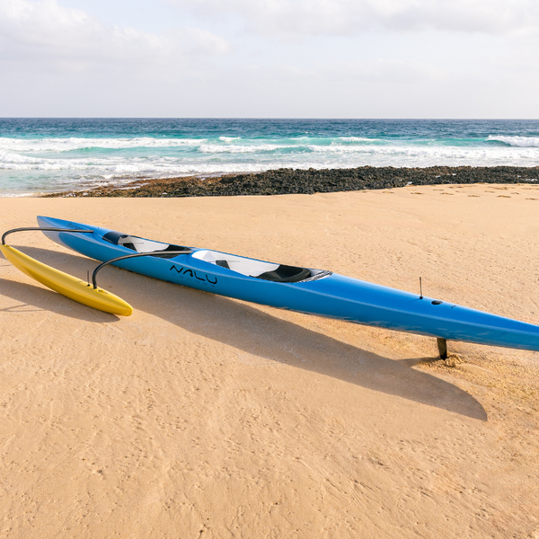 A side profile of a light blue Synergy tandem racing canoe from the Nalu collection on a beach, highlighting its aerodynamic hull, dual cockpits, and high-visibility yellow ama.