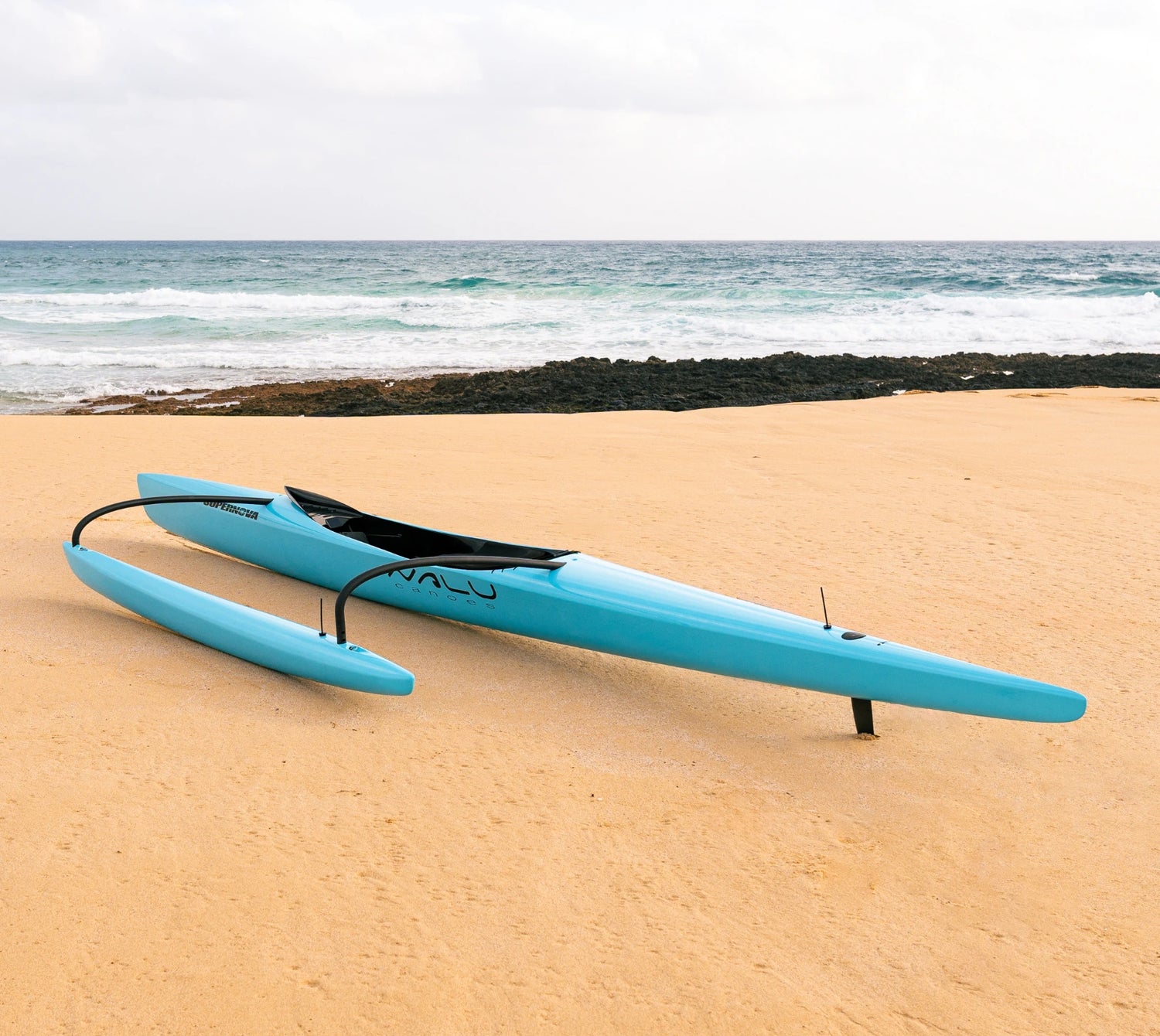 A high-performance teal racing canoe from the Nalu Outrigger collection rests on a sandy beach with crashing ocean waves in the background, showcasing its sleek carbon-fiber construction.