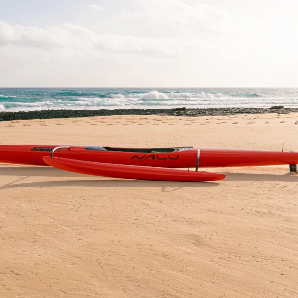 A wide side view of the red Shaka high-performance outrigger canoe on a beach, emphasizing the ama and connecting iakos against the ocean horizon.
