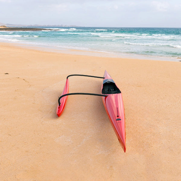 A high-angle view of a sleek red high-performance outrigger canoe from the Nalu Shaka Collection resting on a sandy beach facing the turquoise ocean.