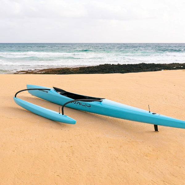A teal Supernova High Performance outrigger canoe positioned on the sand facing the ocean waves, highlighting its sleek hydrodynamic hull.