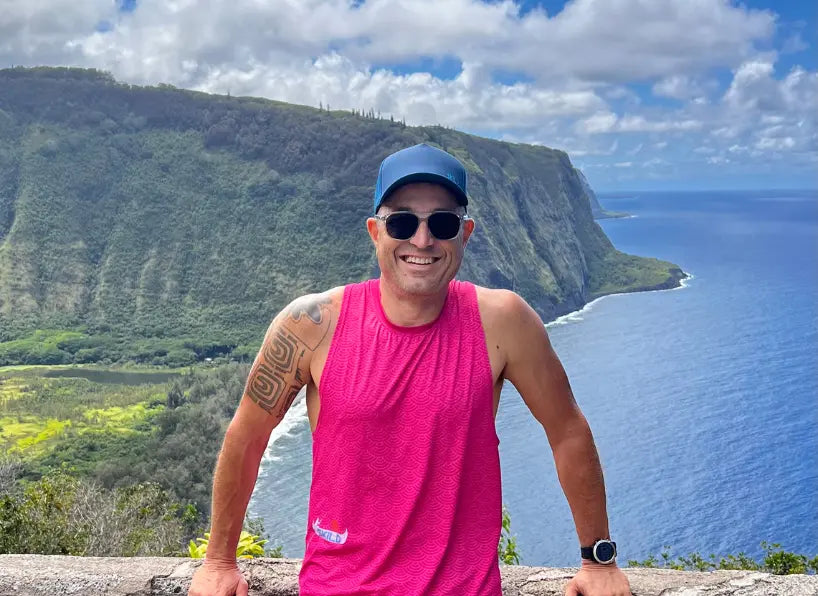 A smiling male athlete with a shoulder tattoo wearing a vibrant pink performance tank top from the Racerback collection, standing against a backdrop of lush green cliffs and a deep blue ocean.