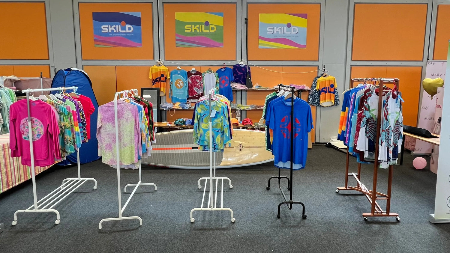 An indoor retail display featuring several clothing racks filled with a wide variety of colorful, patterned performance rashguards from the whole collection, with Skild brand posters and a paddleboard in the background.