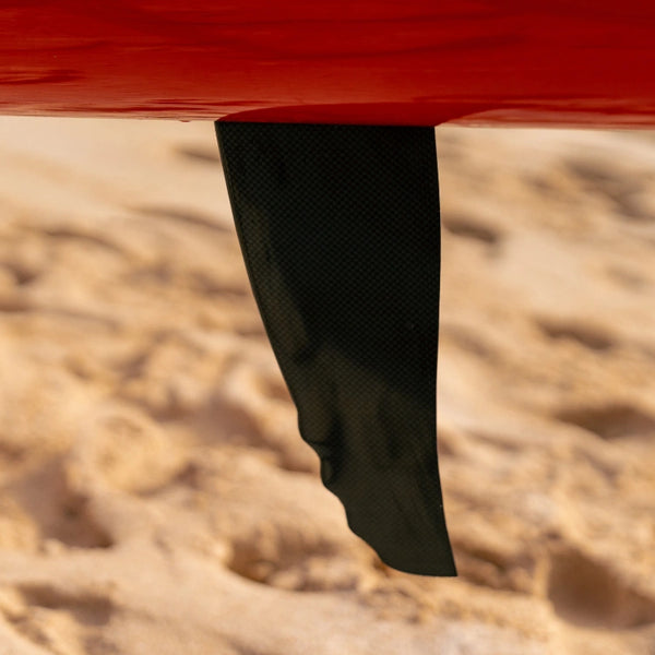 A close-up of the black carbon fiber tracking fin on the underside of a red Shaka high-performance outrigger canoe.