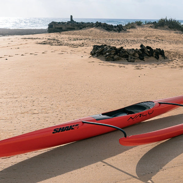 Mid-profile view of a red Shaka high-performance canoe featuring the Nalu and Shaka logos on a beach with volcanic rock formations.