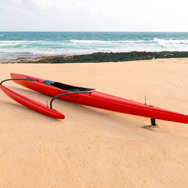 A low-angle side profile of a red Shaka high-performance canoe on a sandy beach, highlighting the sharp hull design and the black tracking fin.