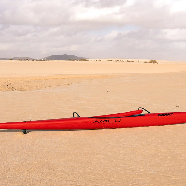 A side profile of the red Shaka high-performance racing canoe on an expansive sandy beach with desert dunes and mountains in the background.