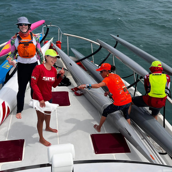 A team of athletes in red and orange performance gear from the Skild collection secures outrigger canoes onto a transport boat during an offshore training session.