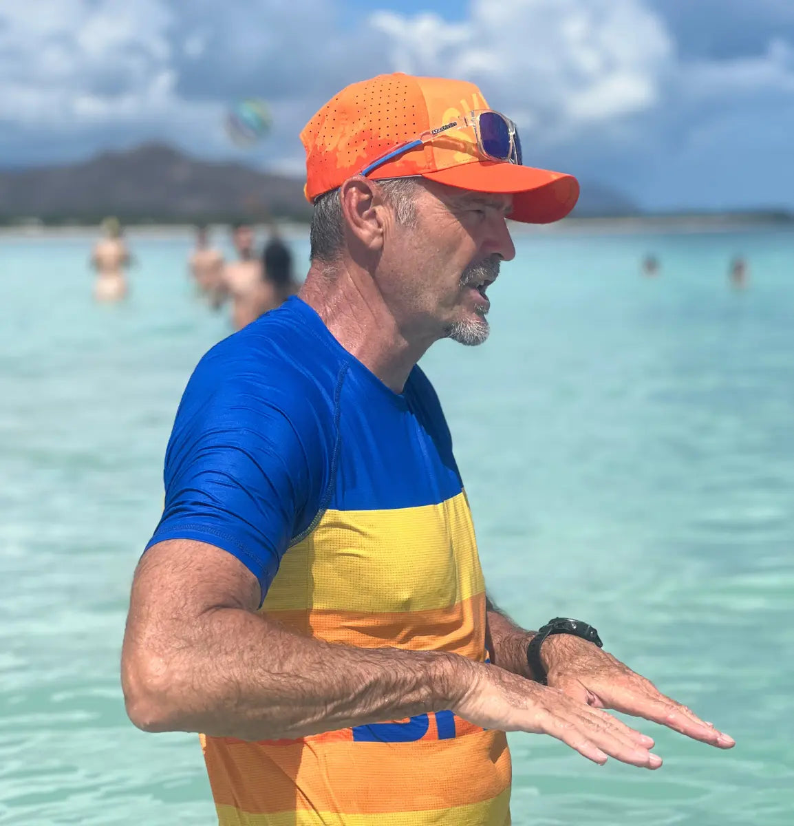 A professional male coach wearing a blue and yellow performance top from the Skild rash guard collection and an orange brand cap gives instructions while standing in clear tropical turquoise water.