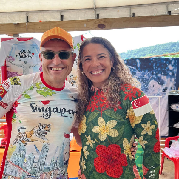 A smiling man in an orange brand cap and a woman in a floral Singapore-themed performance top from the high performance rash guard collection pose together at a water sports event.