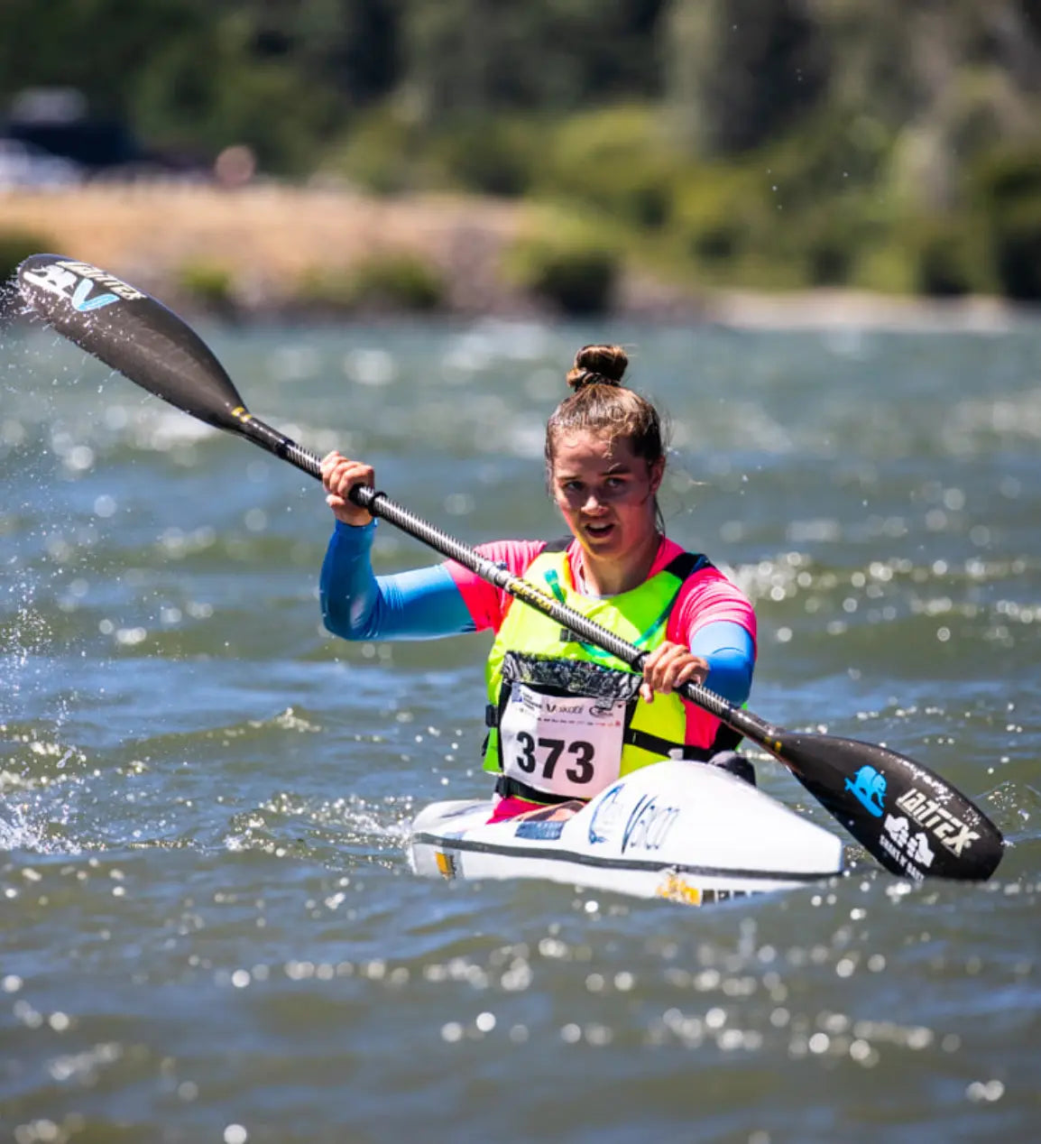 A female athlete wearing a pink and blue performance top from the Skild Champion rash guard collection powerfully paddles her kayak during a high-speed water sports competition.