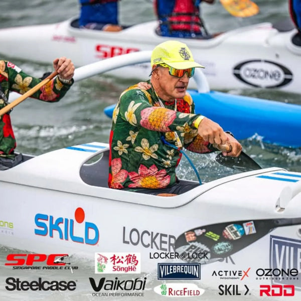 An elite male athlete wearing a green and red floral high performance rash guard from the Skild collection paddles a white outrigger canoe during a competitive race.