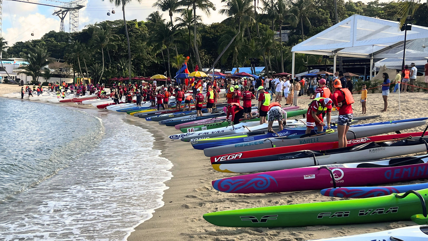 A wide panoramic view of a paddle competition on a tropical beach with a long lineup of colorful outrigger canoes and athletes preparing for a race under a bright sky.
