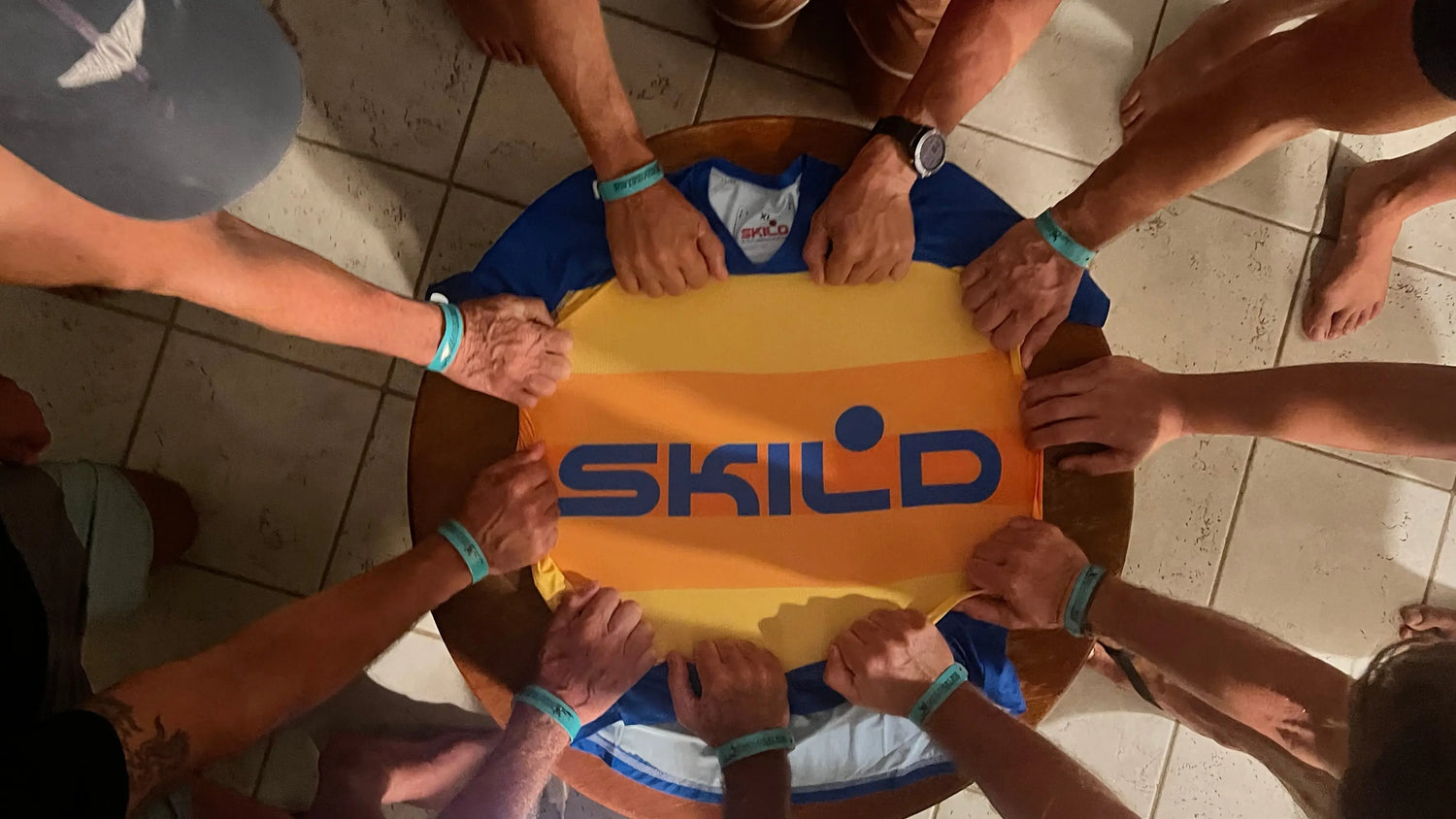 Multiple athletes gather around a table, placing their hands on a vibrant Skild performance jersey from the yellow collection to showcase team unity and branding.