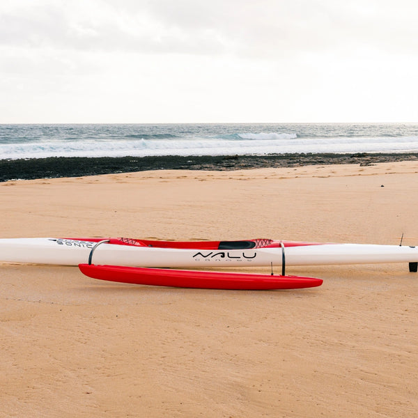 A full-length side profile shot of the Nalu Supersonic outrigger canoe on the sand, highlighting the red ama and aerodynamic hull.