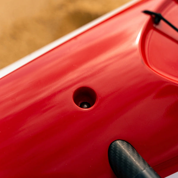 A recessed drainage valve on the red hull of a Nalu outrigger canoe designed for efficient water expulsion during performance use.