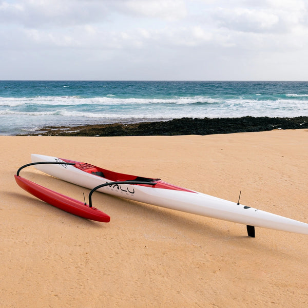 Side perspective of a sleek white and red outrigger canoe on a tropical beach facing the blue ocean waves.