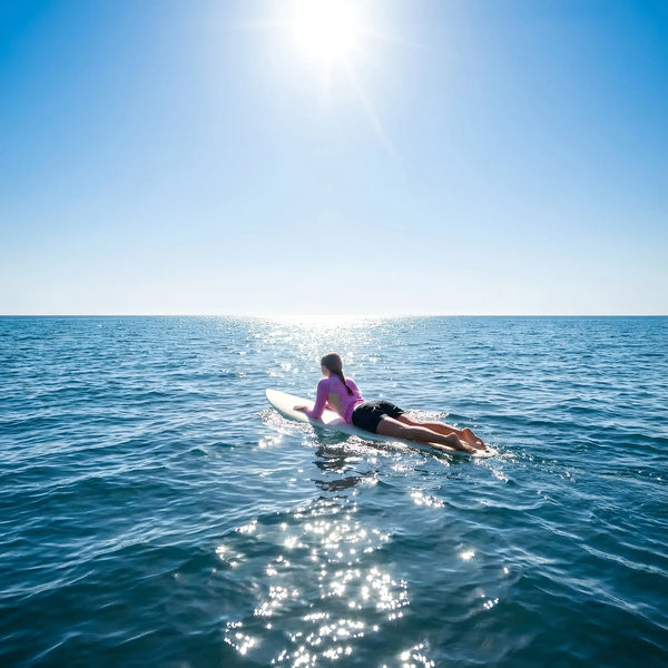 A woman in a pink long-sleeve performance top from the Womens Rash Guard collection lies on a surfboard, paddling across sparkling blue ocean water under a bright midday sun.